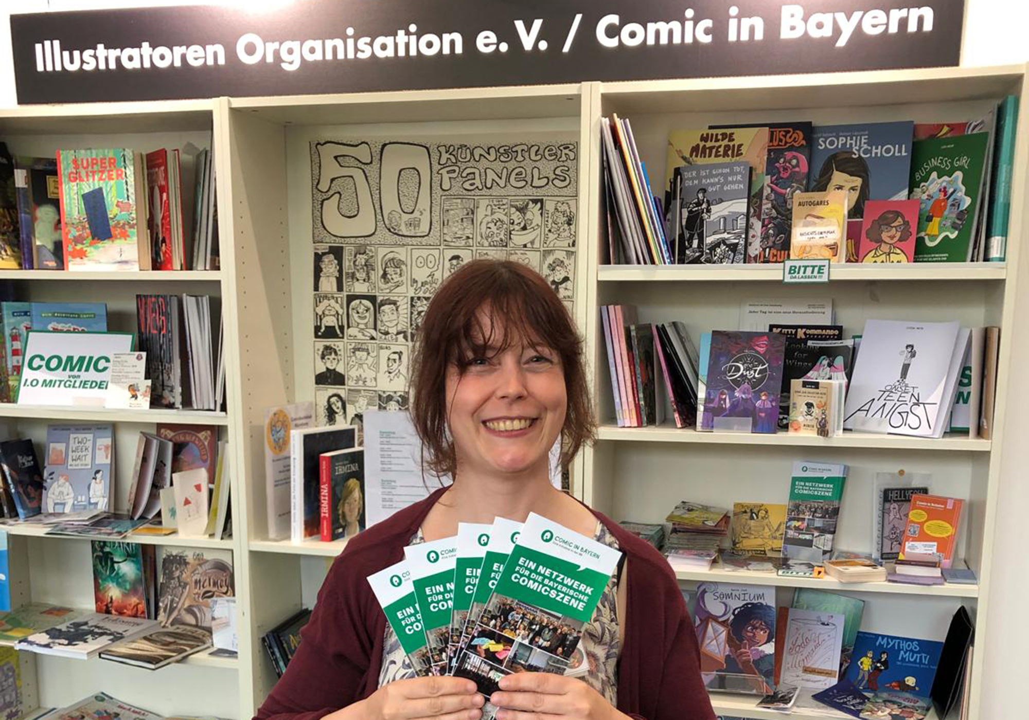 Eine Frau hält vor einem Comicregal lächelnd Flyer von Comic in Bayern hoch, darüber das Schild „Illustratoren Organisation e. V. / Comic in Bayern“.