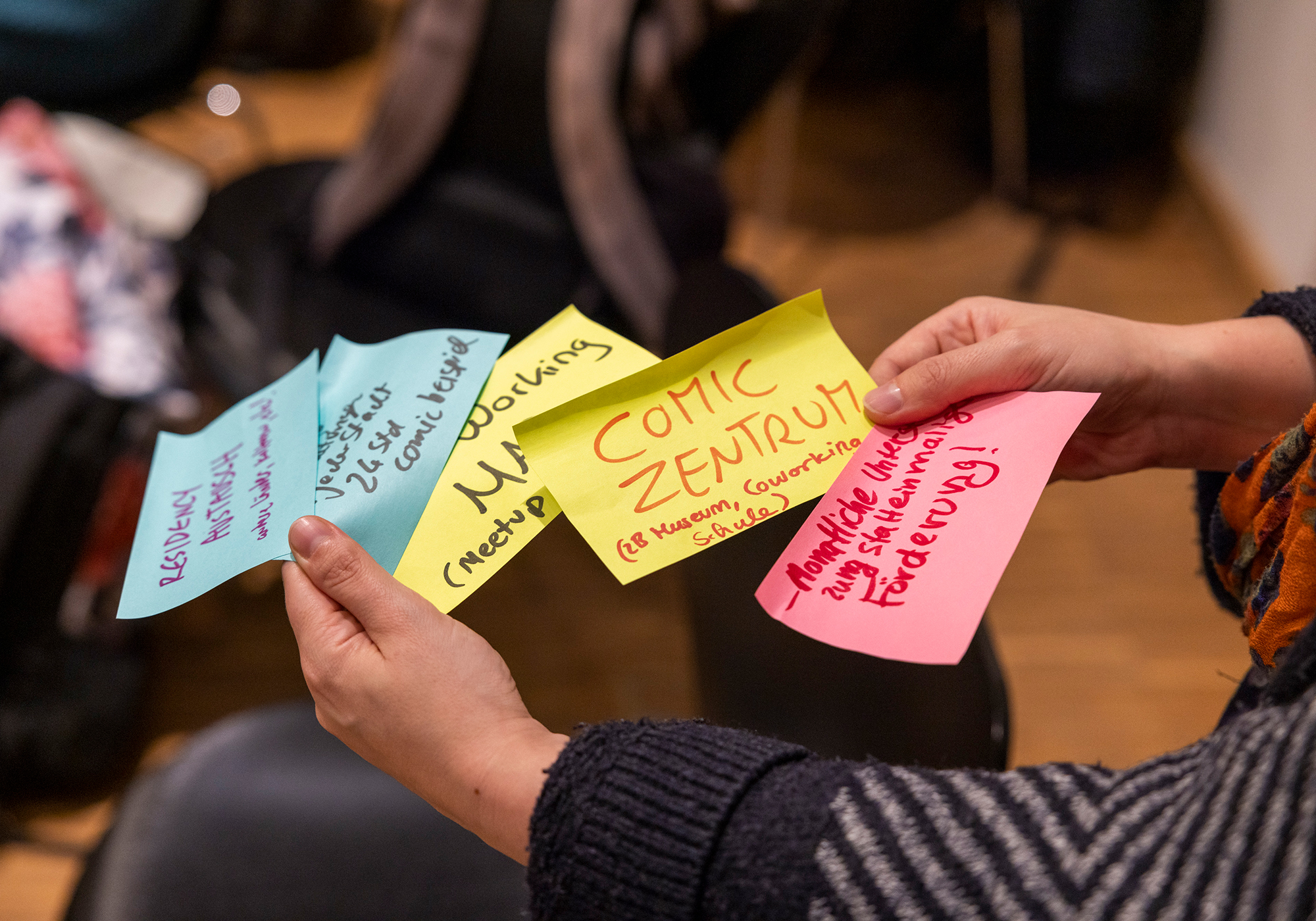 Eine Hand hält bunte Notizzettel mit Ideen wie „Comic-Zentrum“, „Residenz“ und „Förderung“.